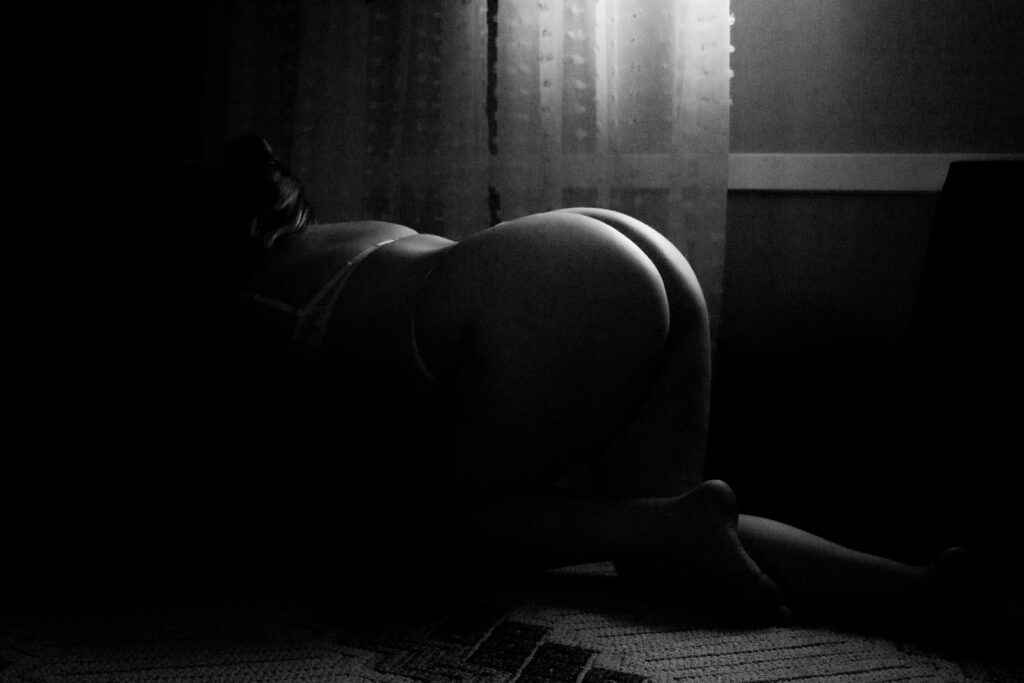 Dark and moody black and white image of a woman with her knees on the floor and her arms on a bed, taken from the floor to silhouette her body in lingerie