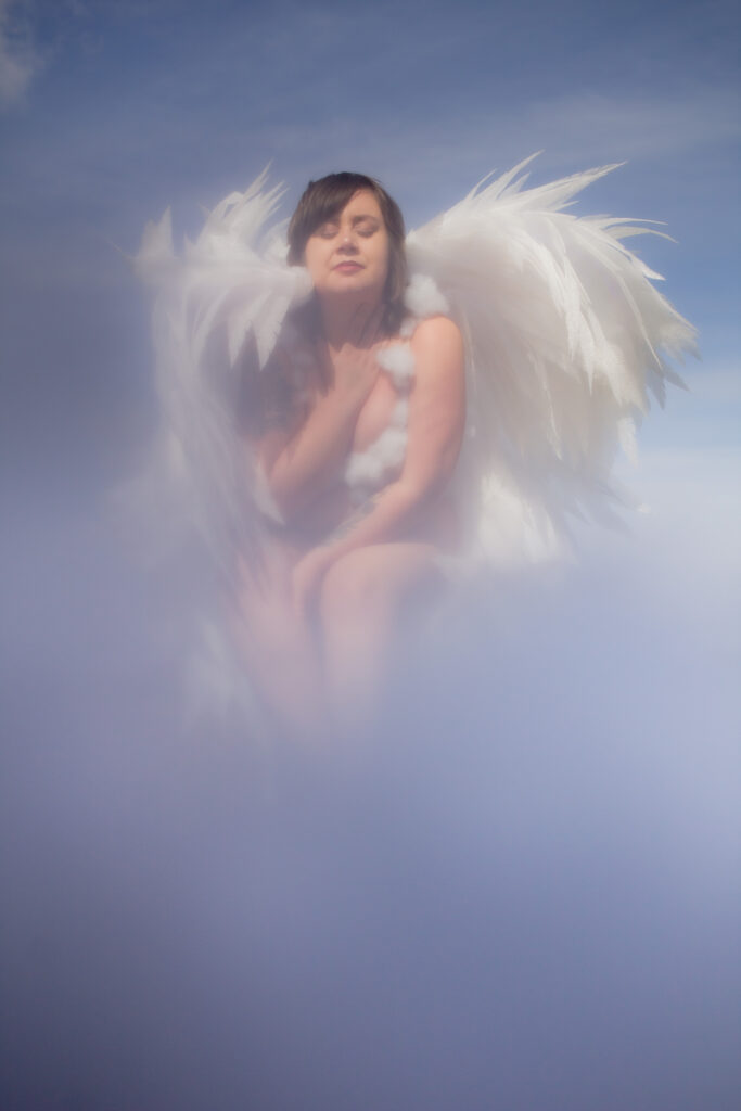 Boudoir photo of a sky angel wearing cloud lingerie and with white wings as she appears to sit in the clouds