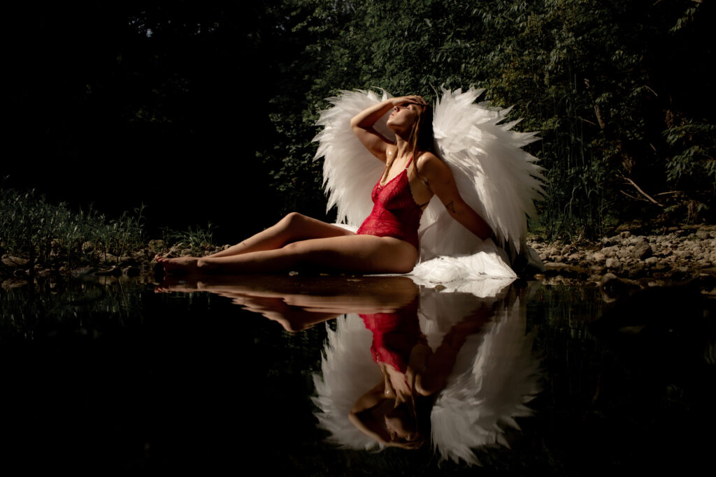 A boudoir photo of a winged baddie lounging by the river with her reflection doubling her in the water below.